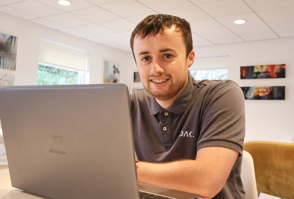 DAK Digital Founder, Ryan Dakin, sat at a desk with his HP Z Book laptop in front of him, Ryan is wearing a grey polo shirt with the DAK Digital logo printed on.